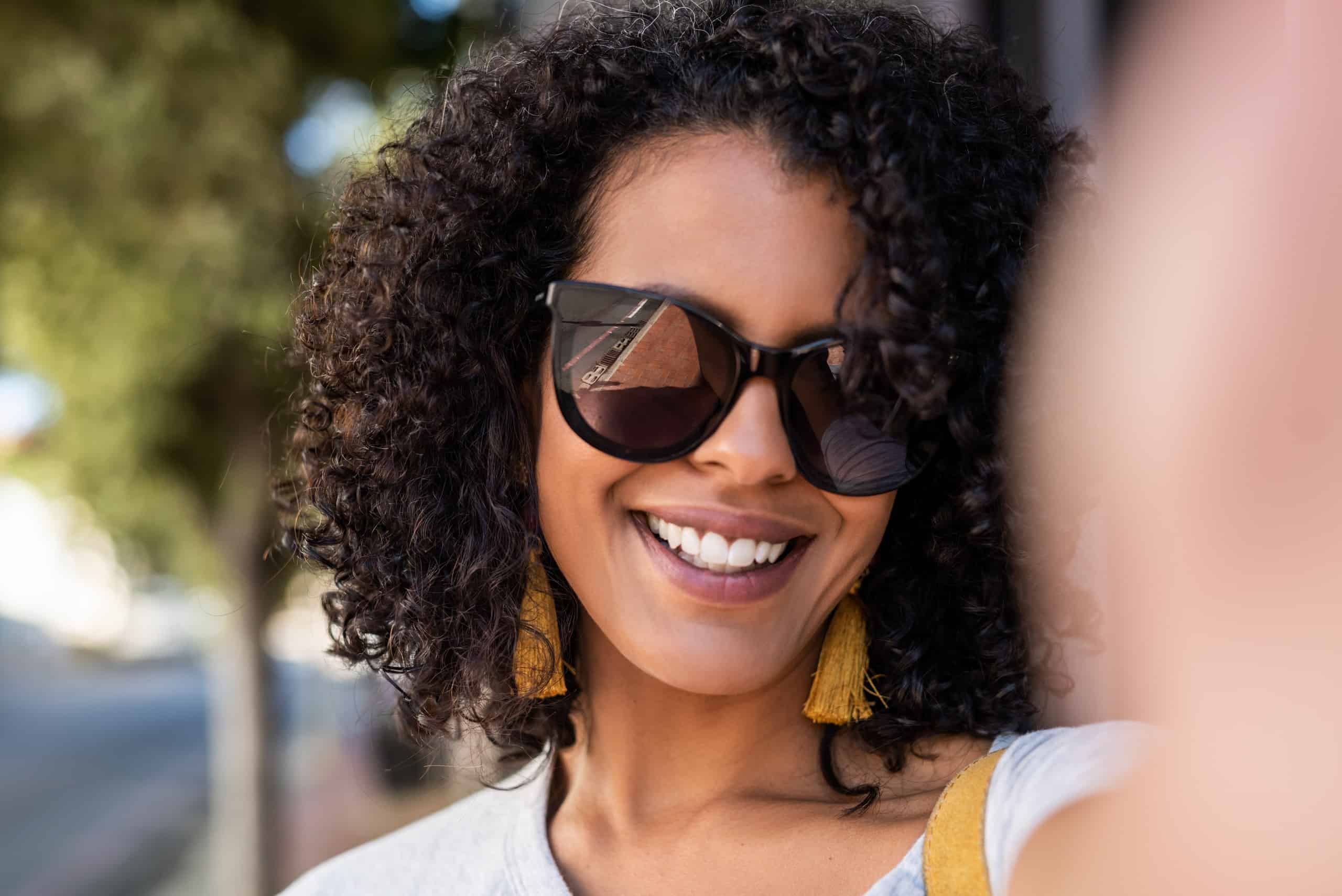 Smiling young woman with curly hair and wearing sunglasses taking a selfie while standing on a sidewalk in the city